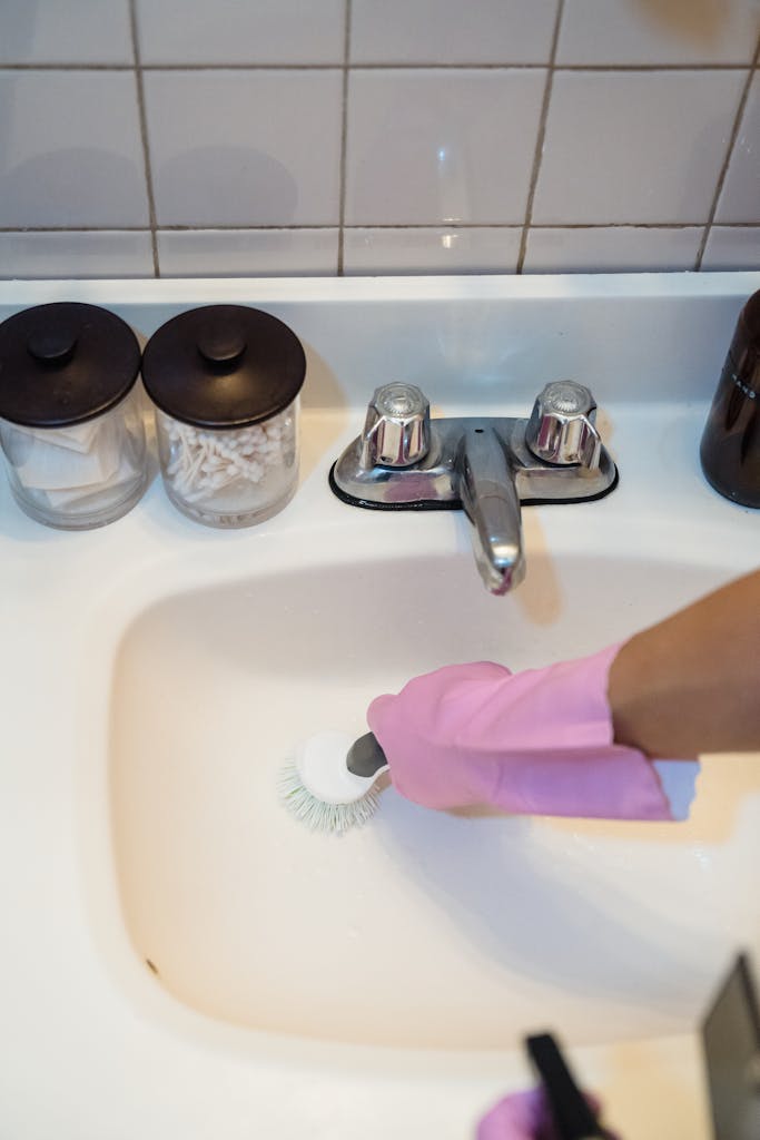 A person using a brush and pink gloves to clean a white bathroom sink, promoting hygiene and cleanliness.
